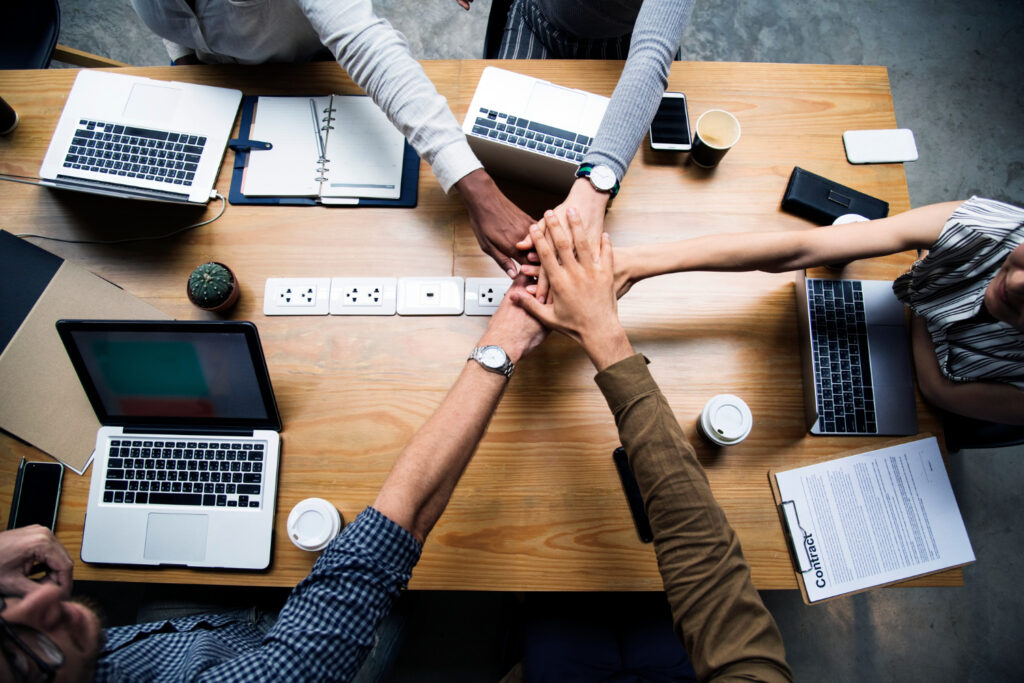 The image shows five team members placing their hands together in the middle of a table, demonstrating team collaboration